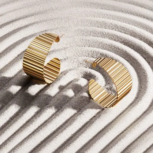 צילום תכשיטים מקצועי של זוג עגילי זהב מונחים על משטח חול מחוספס, משדרים אלגנטיות מודרנית