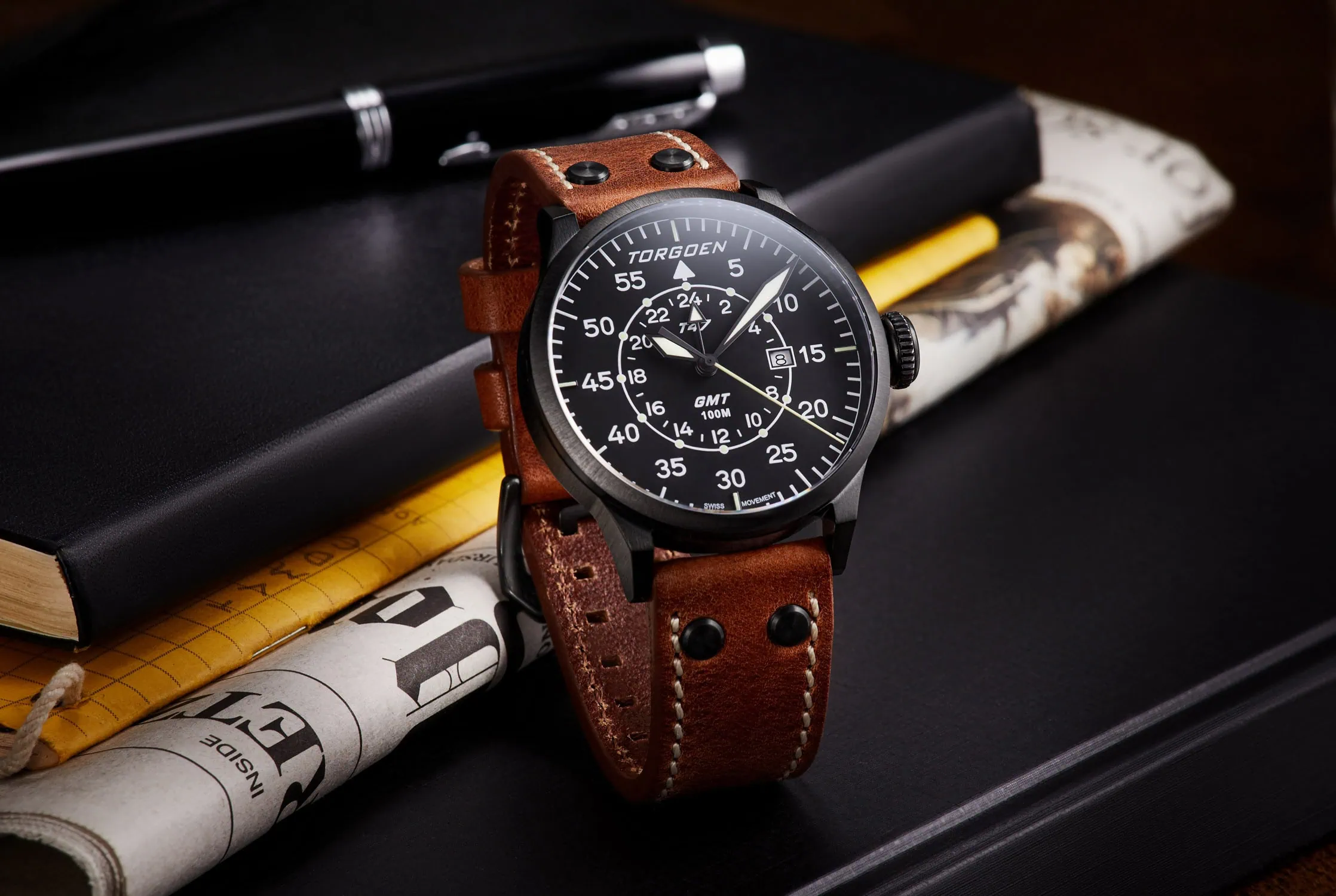 שעון עם לוח שחור אלגנטי ורצועת עור מוצג בין מחברות יוקרתיות כחלק מצילום שעונים לפרסום