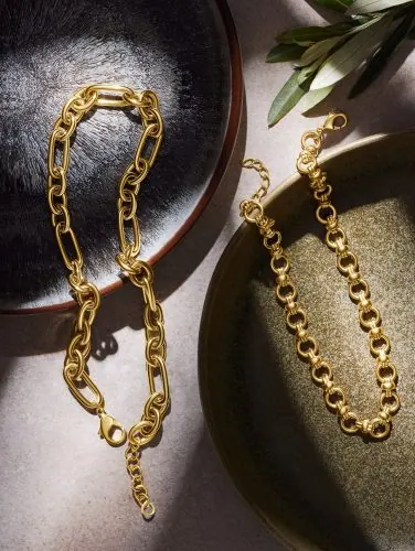 שרשראות זהב מוצגות על צלחות מרקם באור שמש חם, מבטאות יוקרה ונוחות בצילום תכשיטים מקצועי